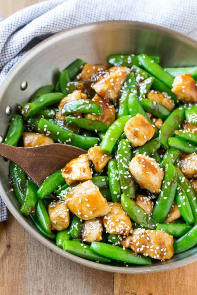 wooden spoon in a pan with cooked chicken bites in sweet sauce along with sugar snap peas