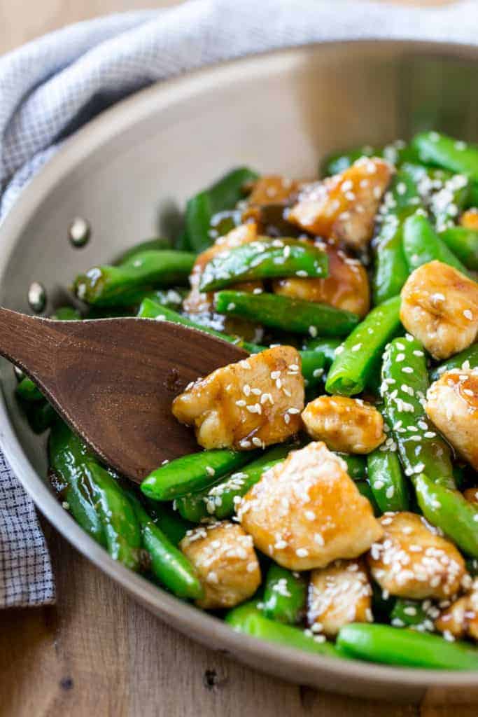 wooden spoon in a pan that has stir-fried chicken bites with green snap peas
