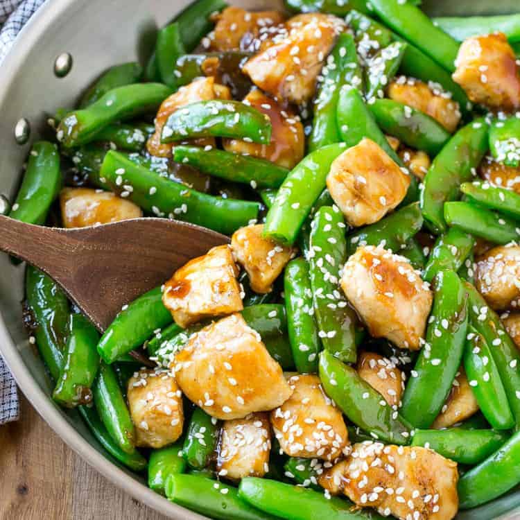 wooden spoon in a stainless steel pan with honey chicken bites stir-fried with sugar snap peas and topped with sesame seeds