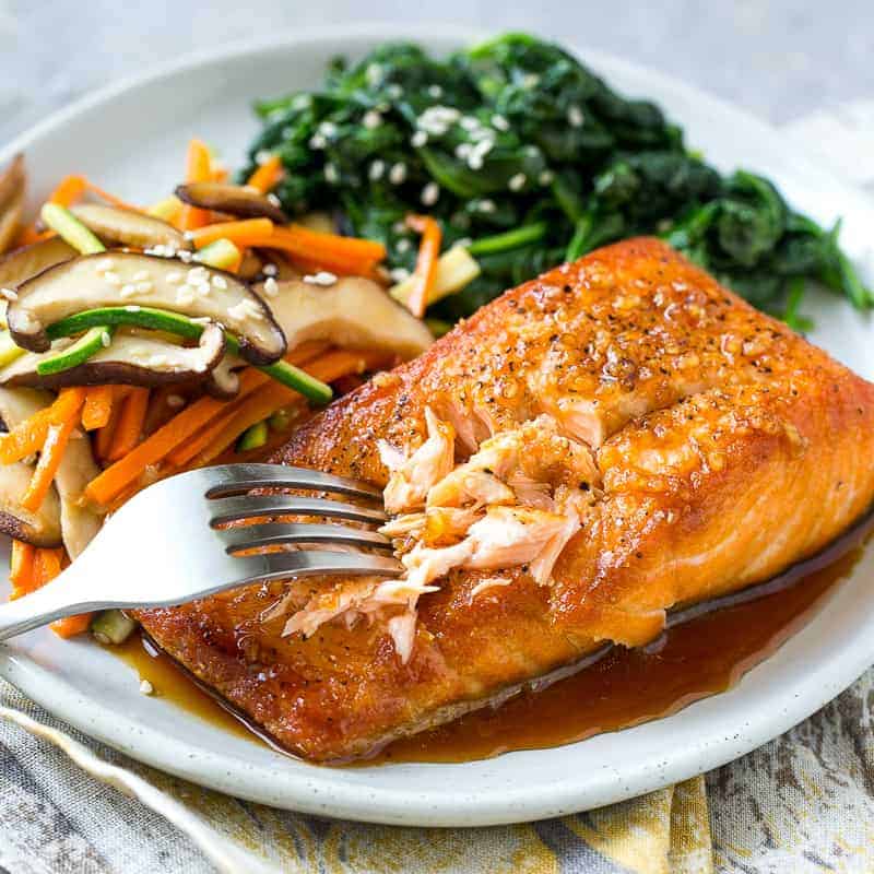 fork flaking a seared salmon on a white plate with a side of sauteed veggies and spinach