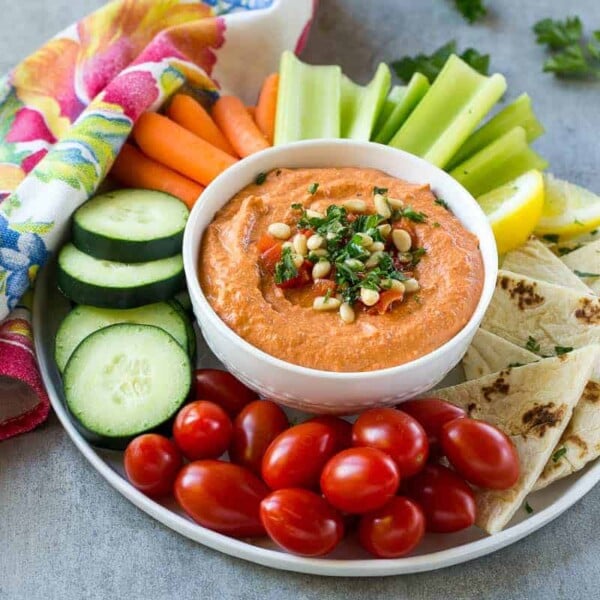 red pepper hummus dip in a small round bowl in the middle of a platter surrounded by veggies.