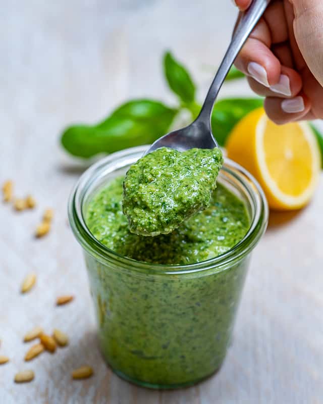 homemade pesto recipe with basil in a jar with a spoonful of pesto and a slice of half lemon behind the jar