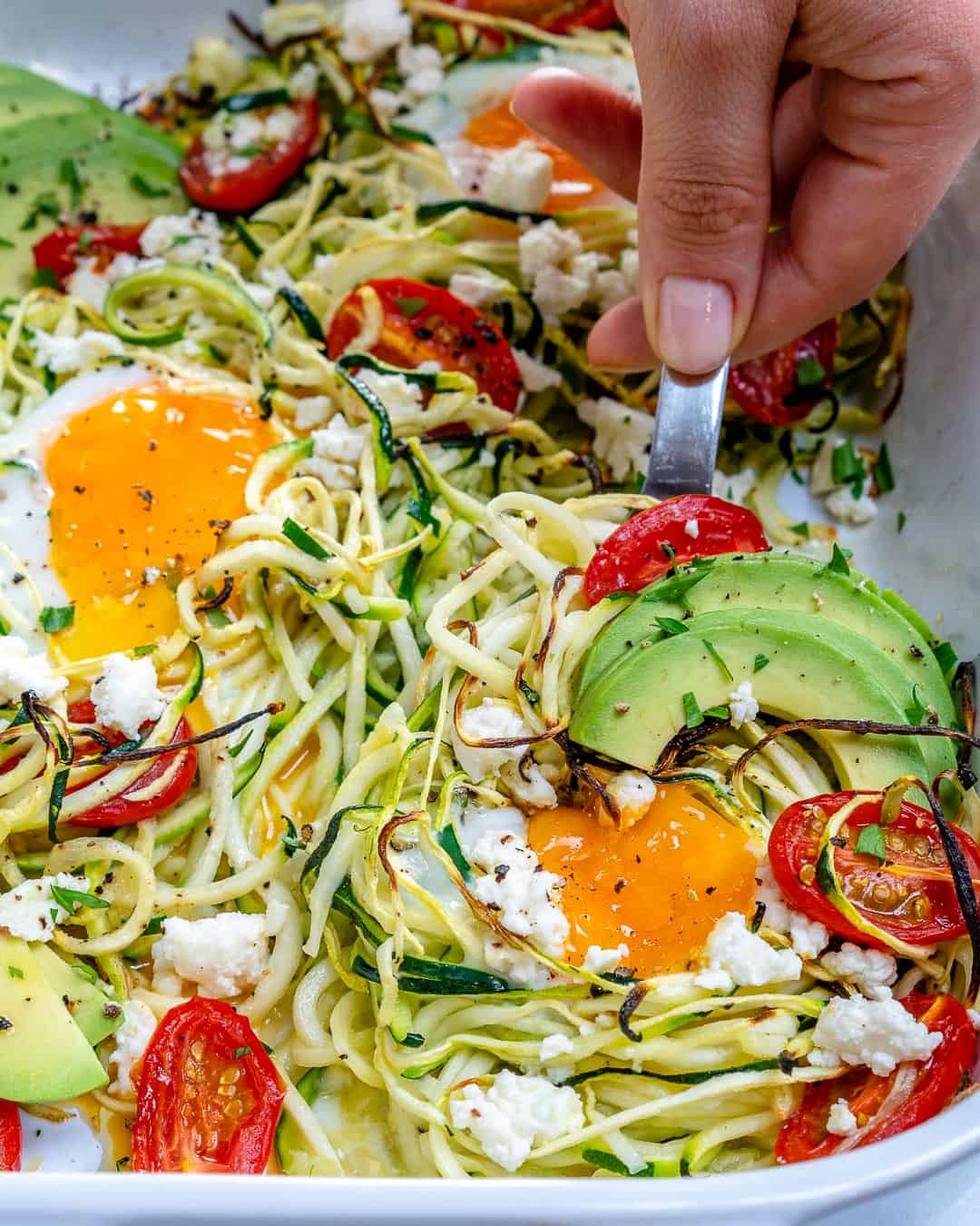 hand grabbing a portion of baked eggs with zoodles in a dish.