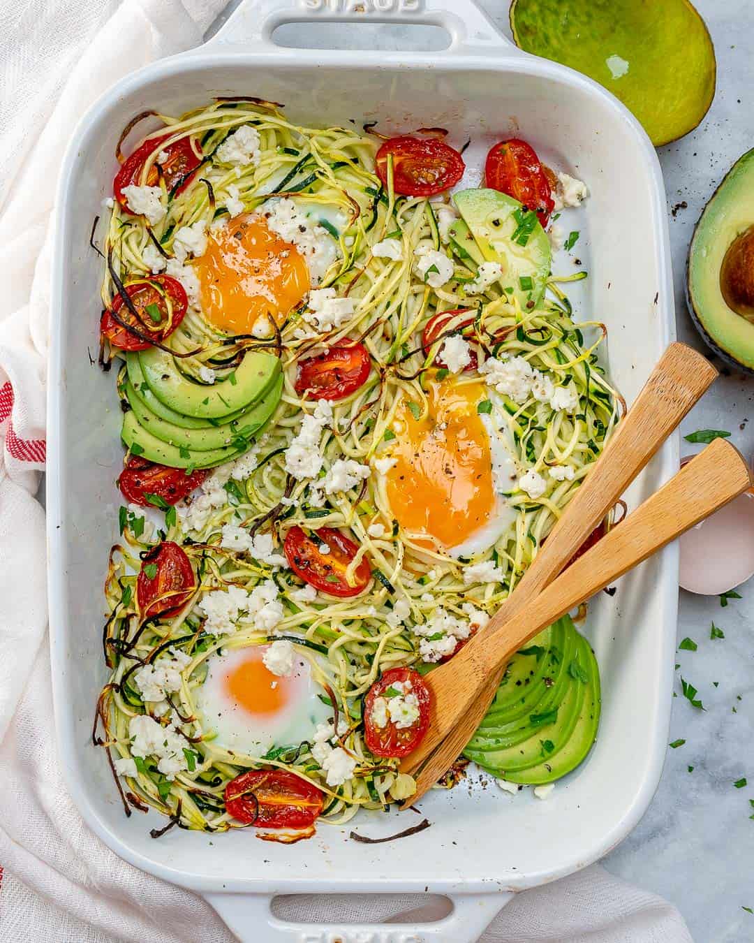 zoodles in a baking dish with baked eggs and cherry tomatoes, topped with sliced avocados.