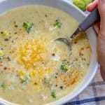 hand holding spoon in a bowl of Broccoli Cheddar Soup recipe topped with shredded cheese