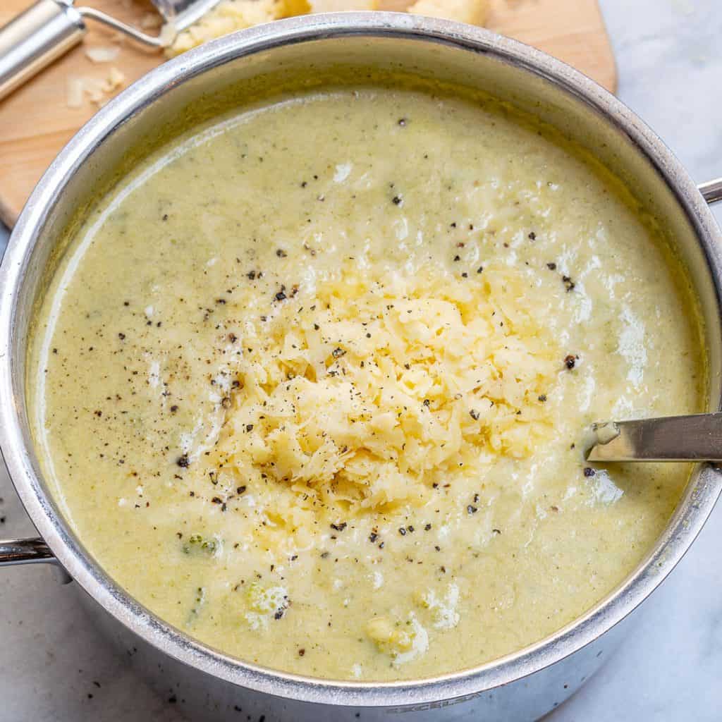 shredded cheese added over creamy broccoli soup in a pot with spoon in pot