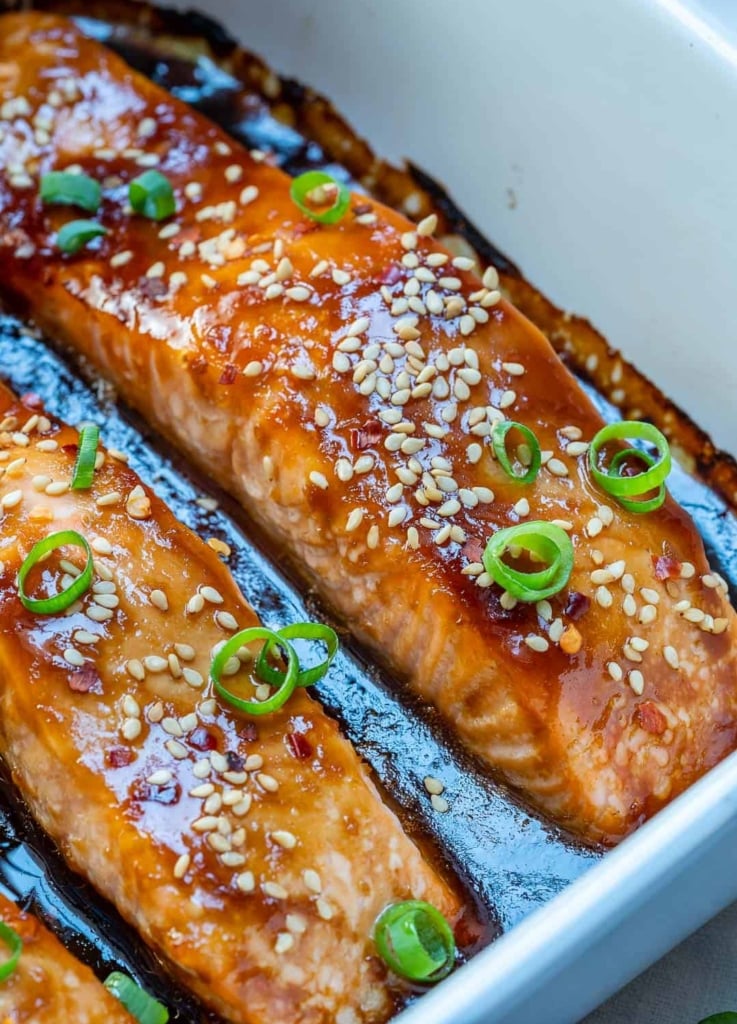 side shot of close up baked teriyaki salmon in a dish with sauce at the botton and the salmon is granished with sliced onions and sesame seeds