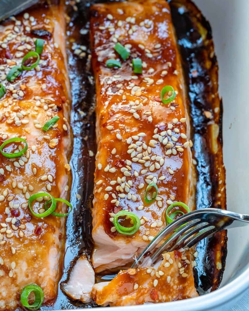 fork flaking a piece of the baked teriyaki salmon filet that is garnished with sesame seeds and sliced green onions