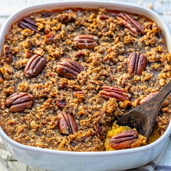 side shot of Butternut squash casserole in a white dish topped with sugar pecan and a wooden spoon in dish