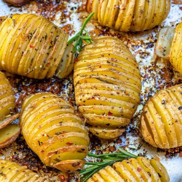 side shot of baked hasselback potatoes on a sheet pan with green rosemary to garnish