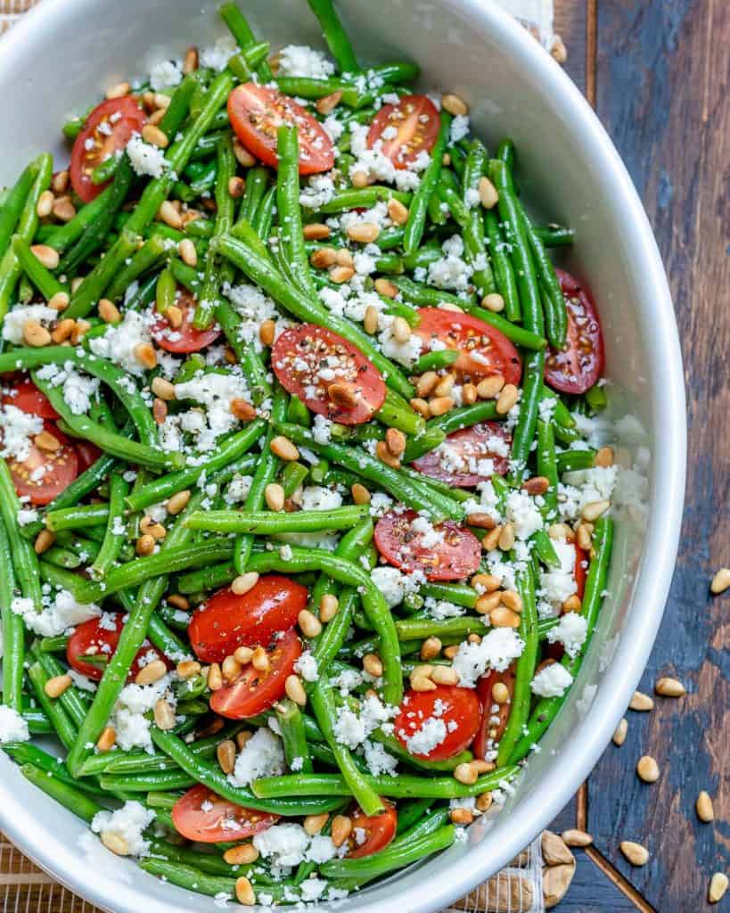 top view of green bean salad in a white bowl with feta cheese and cherry tomatoes, topped with toasted pine nuts 