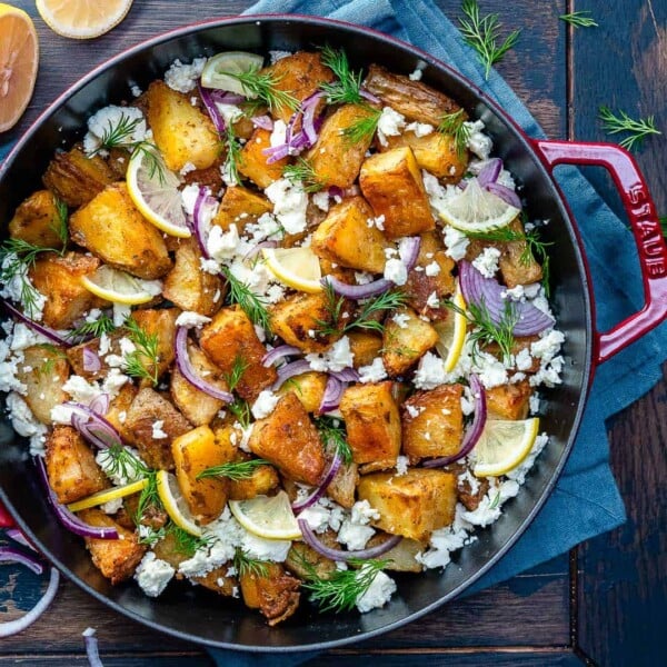 top view of a red skillet with crispy potatoes topped with crumbled feta, fresh dill, lemon garnishes, and sliced red onions