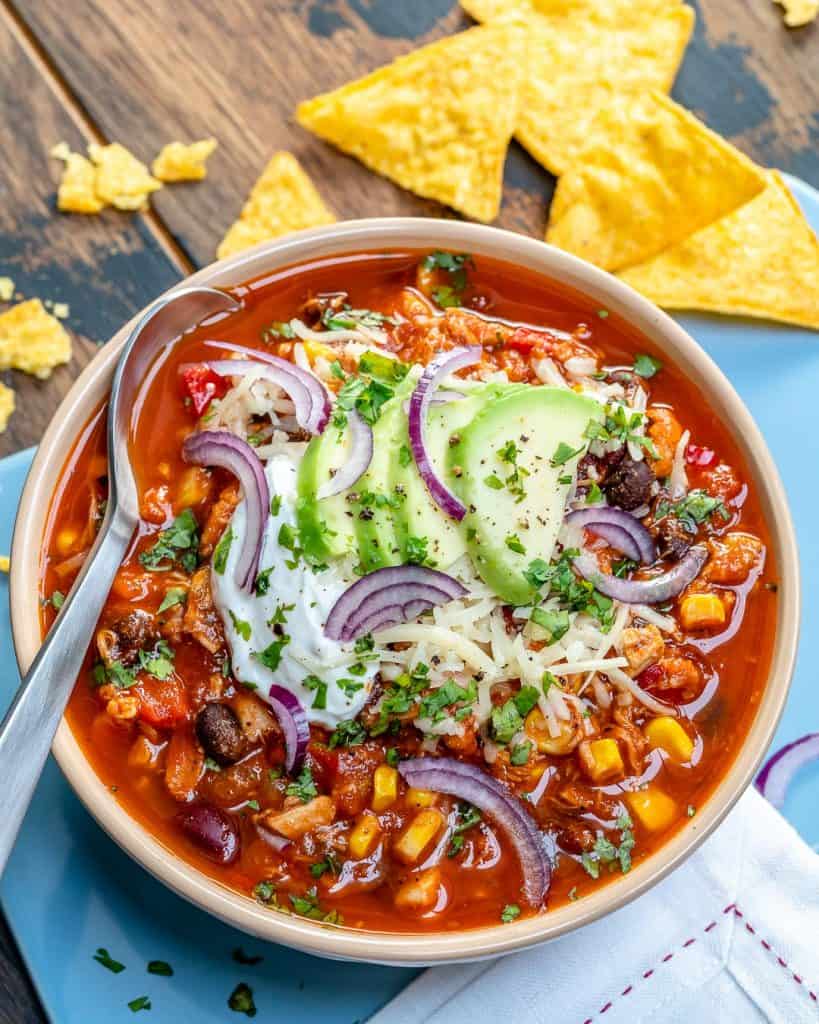 a white round bowl that has red chicken chili topped with shredded cheese, sliced purple onions, avocado, and sour cream