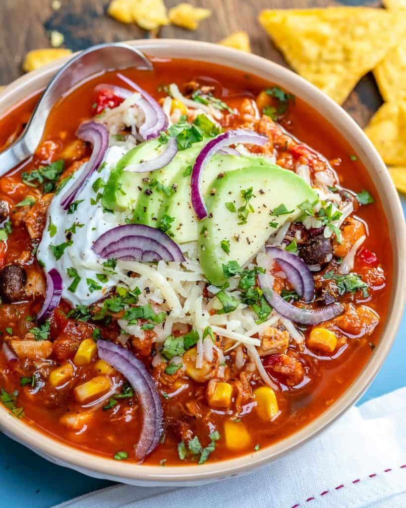 a bowl of chili soup that's red in color made with black beans, chiken, corn, and garnished with shredded cheese avocado, and sour cream