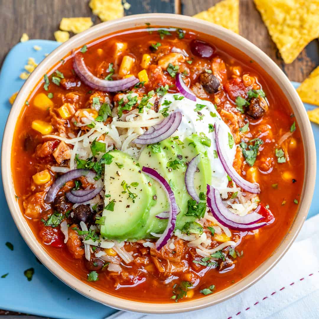 square image of a bowl of chicken chili and is red in color and garnished with shredded cheese, avocado and sour cream