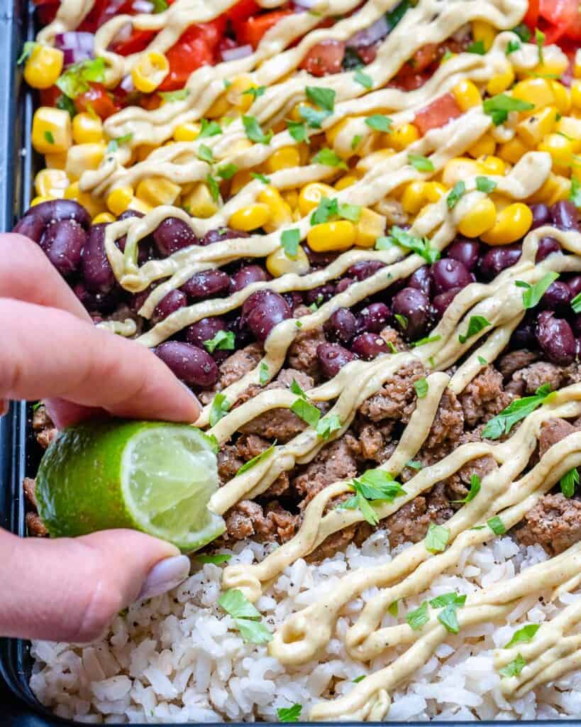 close up shot of a beef burrito bowl with layer of cream sauce and a hand holding a lime wedge squeezing lime into the bowl