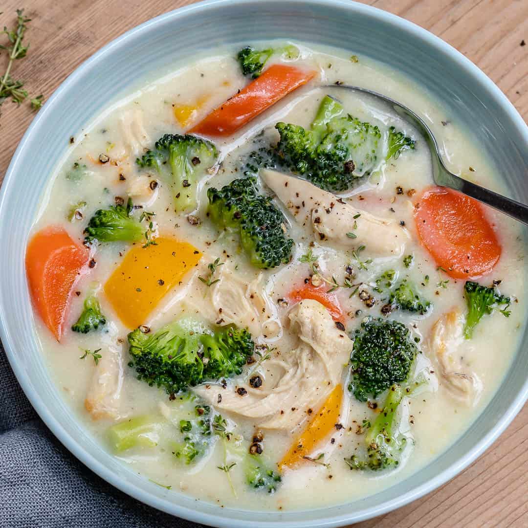 round white bowl of chicken broccoli soup that is creamy looking with yellow bell peppers, and sliced carrots