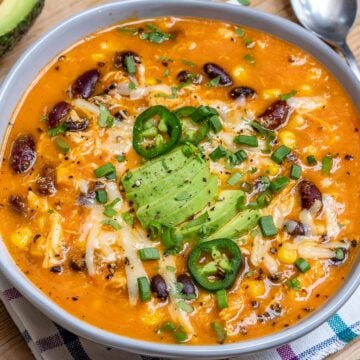side view of chicken enchilada soup in a round bowl topped with shredded cheese, sliced avocado, and sliced jalapenos