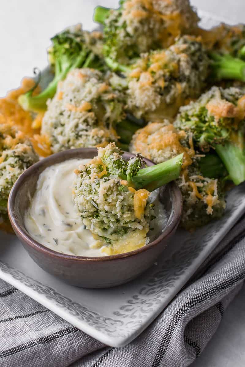 side shot of baked broccoli on a serving plate with one floret in dipping sauce bowl 