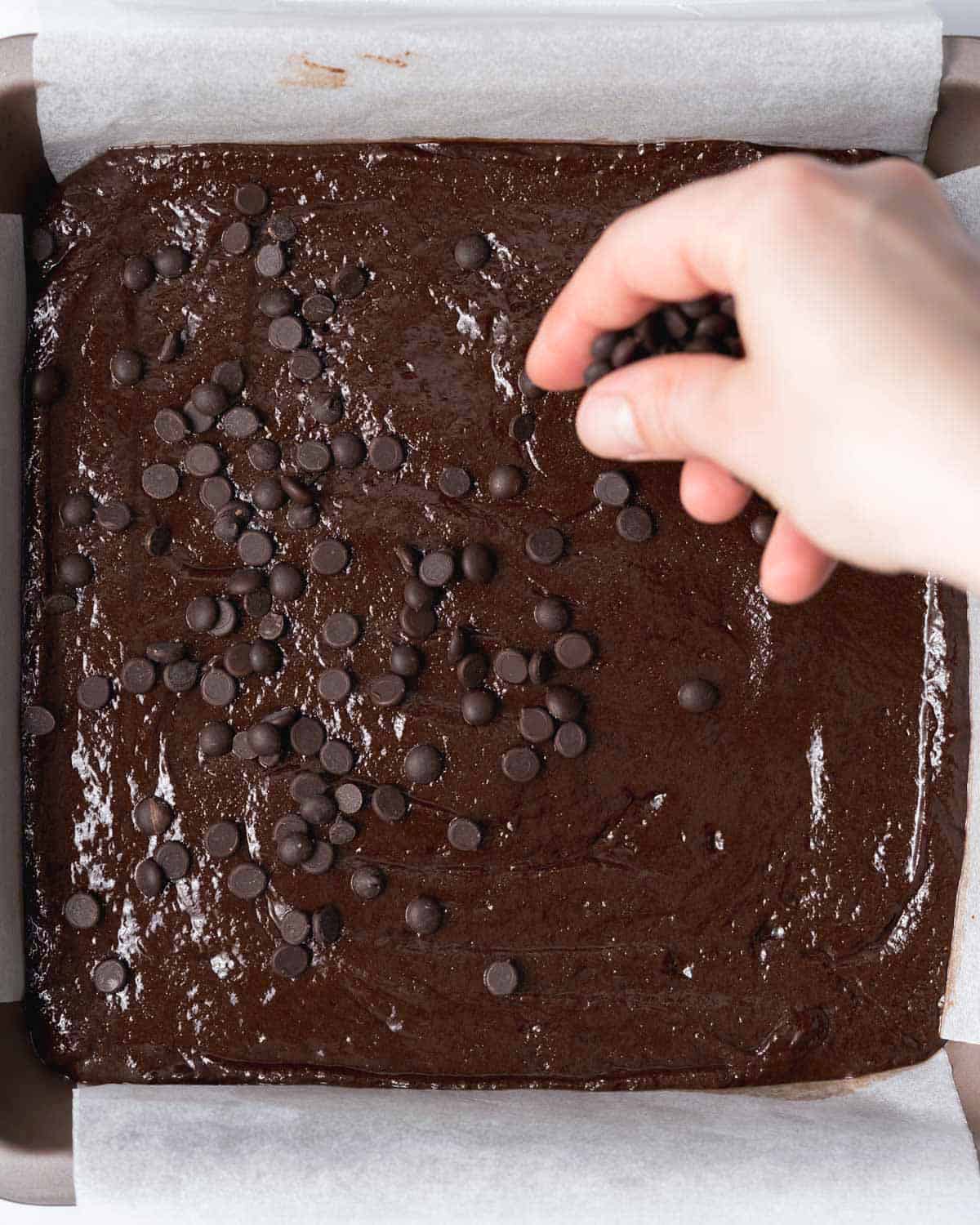 hand adding chocolate chips over brownie batter in a pan.