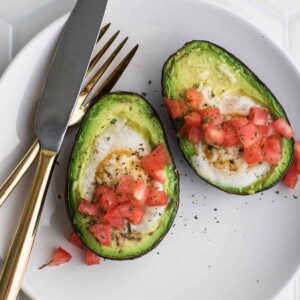 two stuffed avocado with eggs topped with tomatoes on a plate with fork and knife