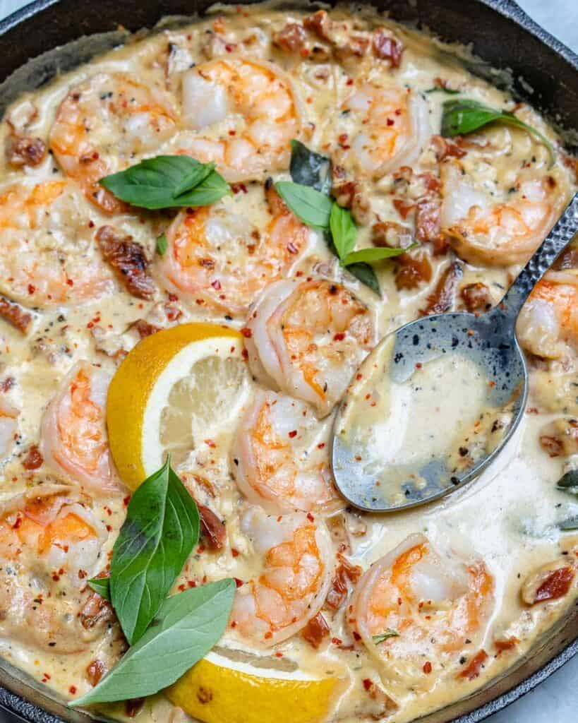 top view of black skillet with creamy shrimp and spoon in skillet, next to skillet on top left there are 2 lemon wedges and top right twi basil leaves
