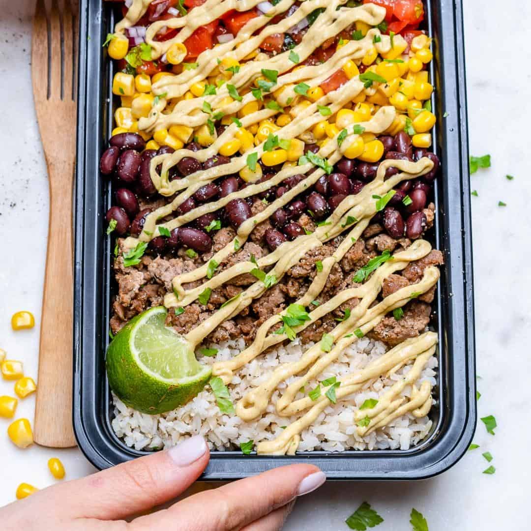 hand holding a black meal prep bowl that has beef burrito, made with brown rice, black beans, corn, ground beef, and salsa. Topped with a creamy sauce