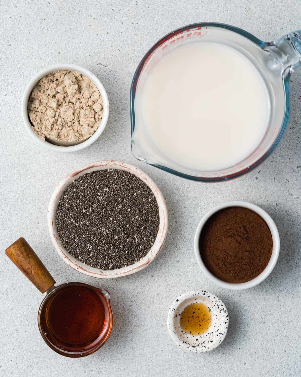 ingredients to make chia pudding: milk, bowl of cocoa powder, maple syrup, chia seeds, vanilla, protein powder.