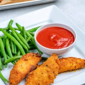 baked chicken strips on a plate with a side of green beans and a small bowl of ketchup.