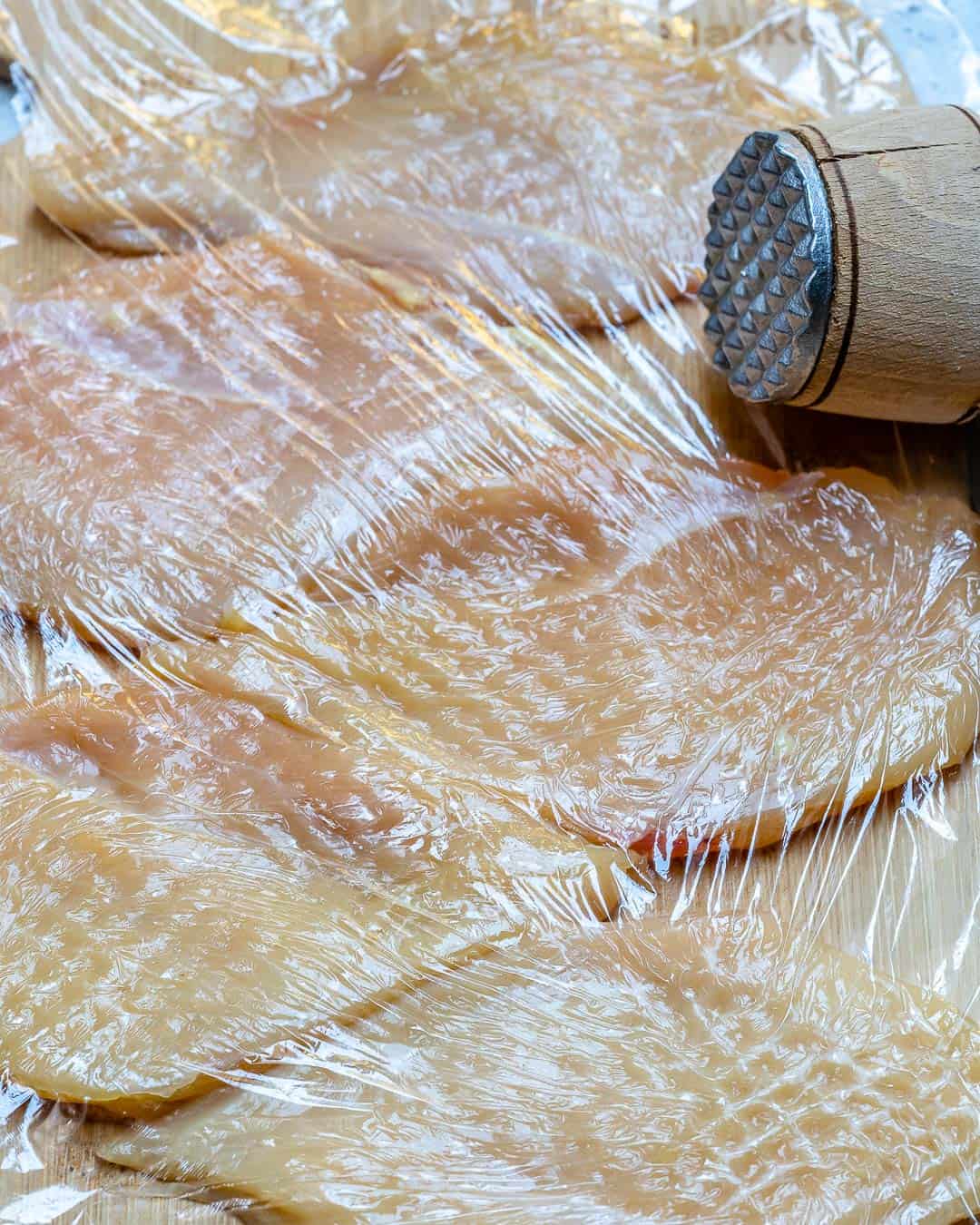 Chicken breast being pounded with a millet covered with plastic wrap.