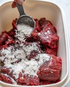 raw beef chunks in a bowl with flour and spices being added over it.