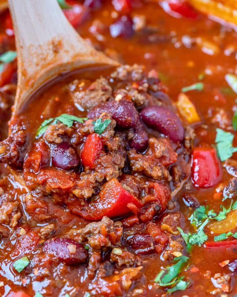 close up a wooden spoon full of chili over healthy beef chili in pot