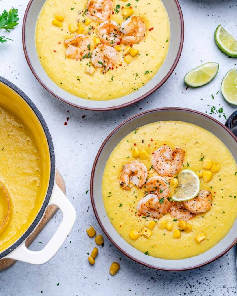 top view of 2 bowls of corn and potato soup thats yellow in color, topped with sauteed shrimp and 3 slices of lime wedges on the side of the soup bowl