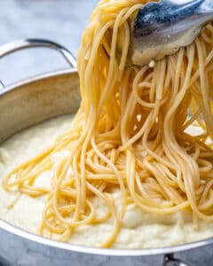 spaghetti being added over alfredo sauce in pan