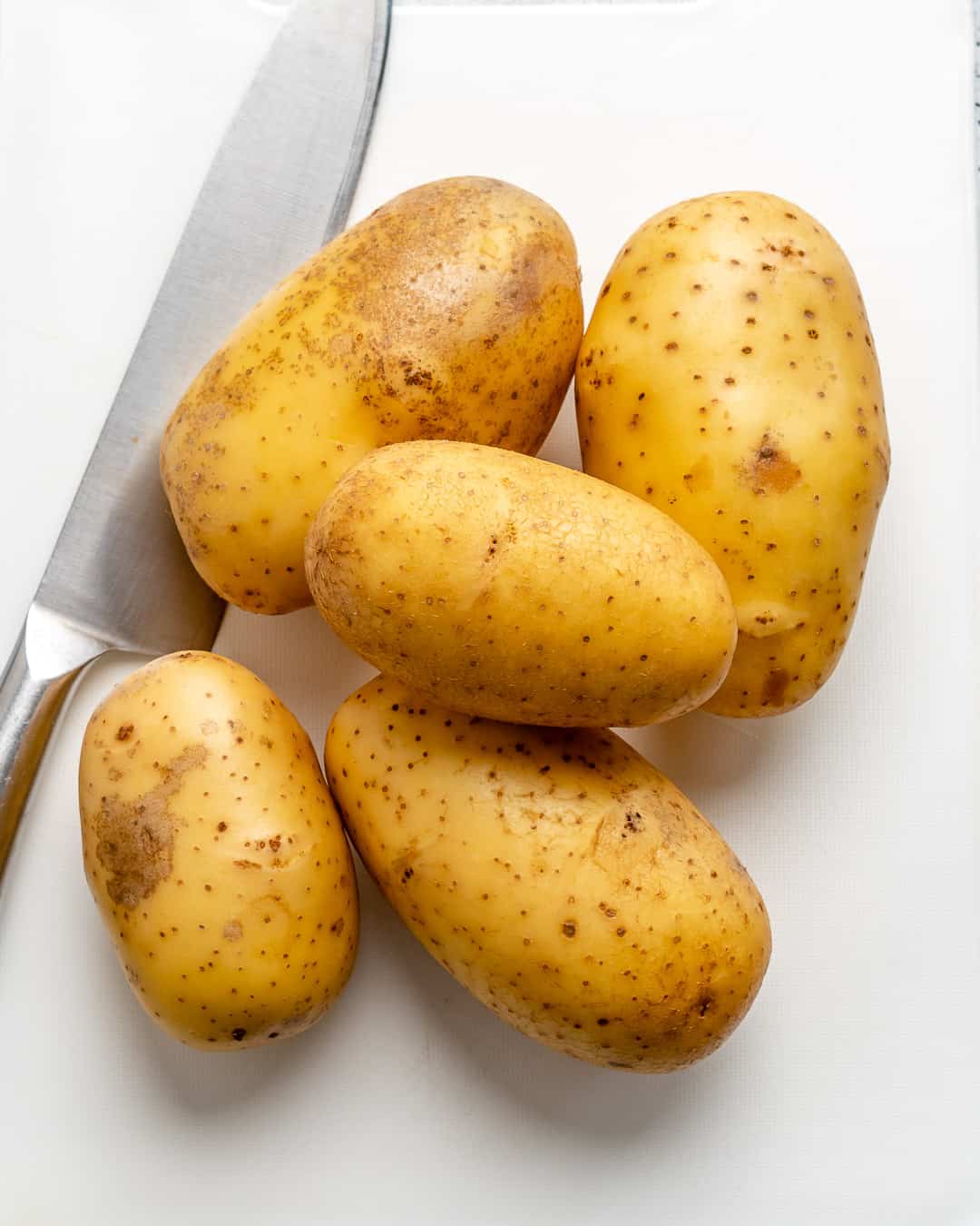 5 yellow potatoes on a cutting board with a knife on the left side.