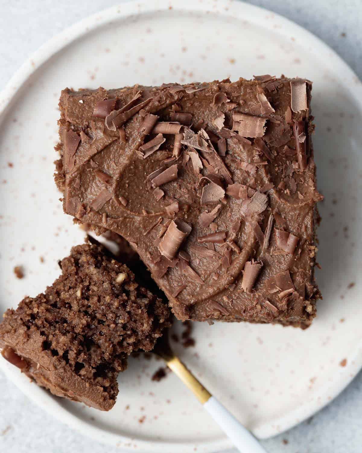 a single square piece of chocolate cake topped with frosting with a spoon cutting a bite off.