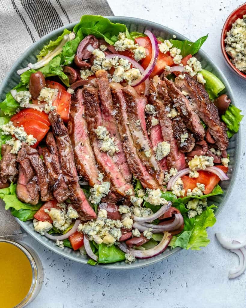 salad in a bowl topped with sliced steaks and  garnishes next to plate