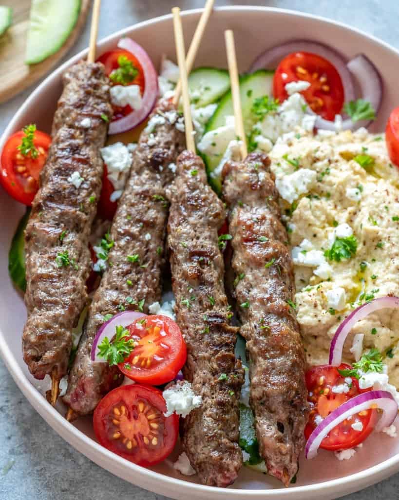side shot of beef kafta kabobs on a plate with hummus and fresh tomatoes 
