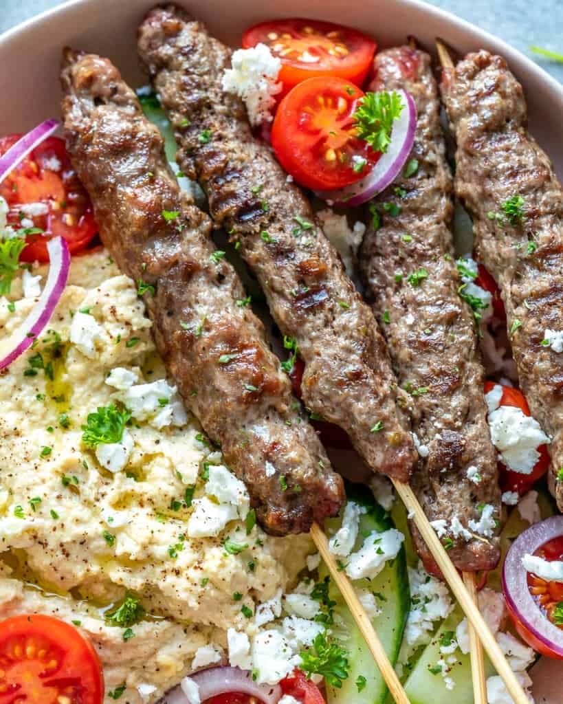 close up view of beef kafta skewers on a plate with tomatoes and hummus 