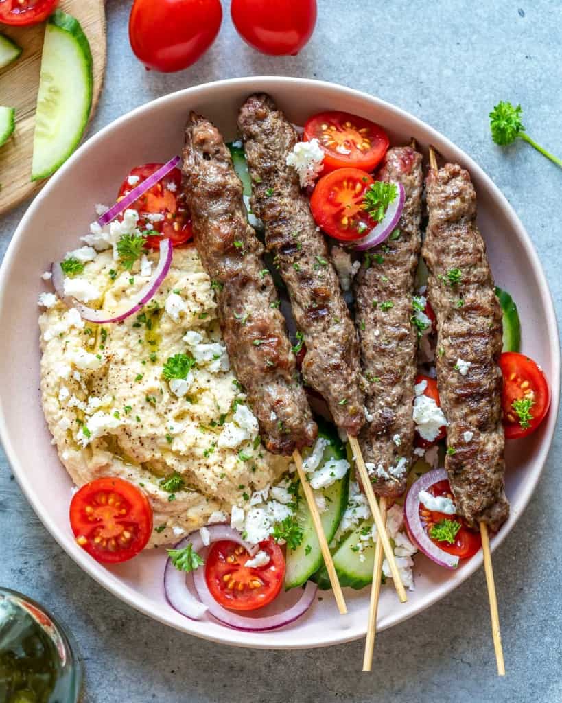 top view of 4 skewers of beef kafta on a round plate  with salad and hummus