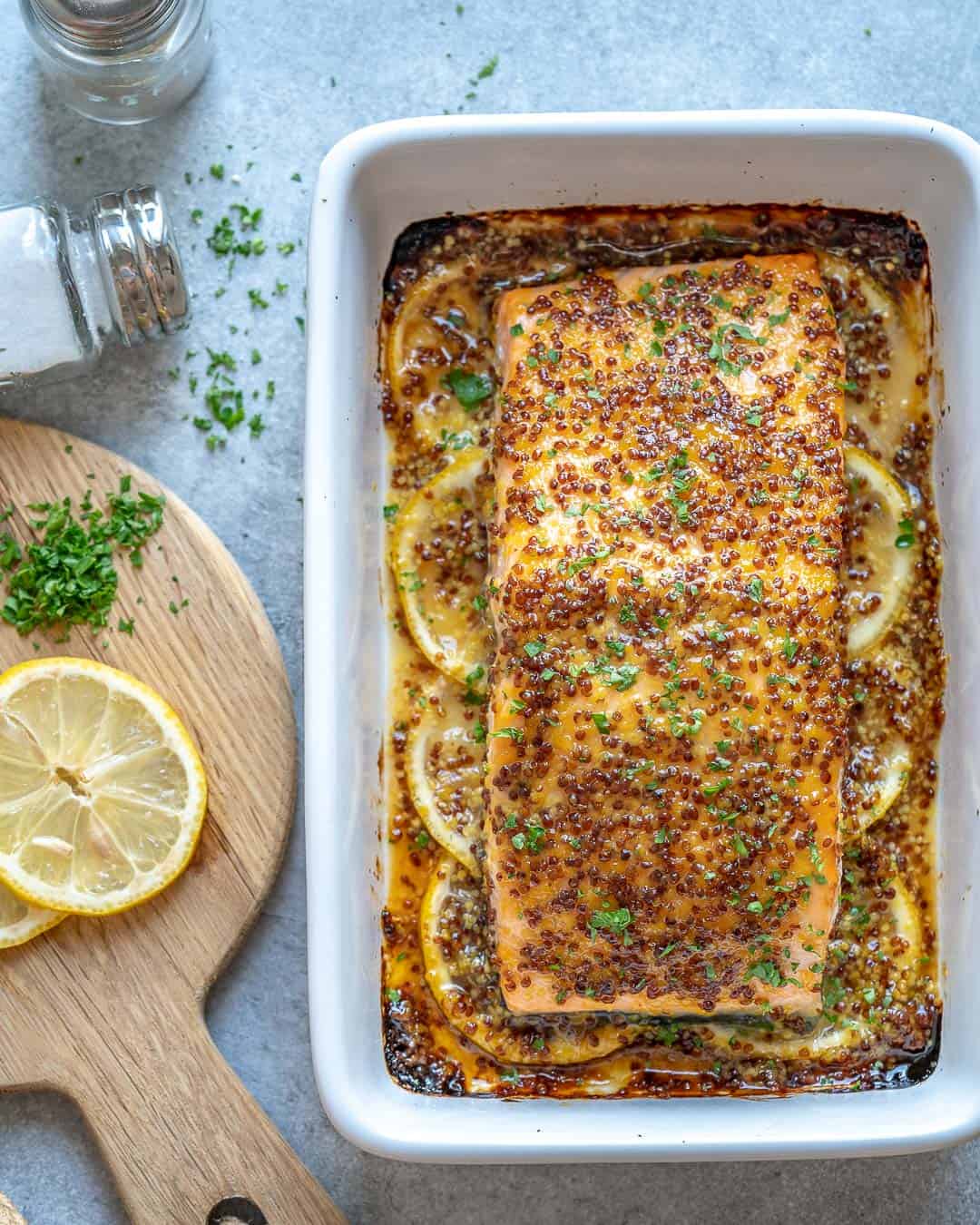 baked salmon fillet topped with honey mustard in a white dish next to a cutting board with fresh lemon slices