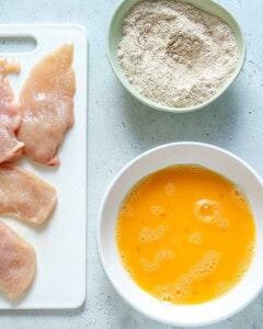 egg wash in a bowl, flour mixture in a bowl, and chicken cutlets on a cutting board,