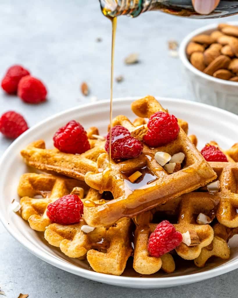 A plate of keto waffles topped with berries with maple syrup being drizzled overtop.