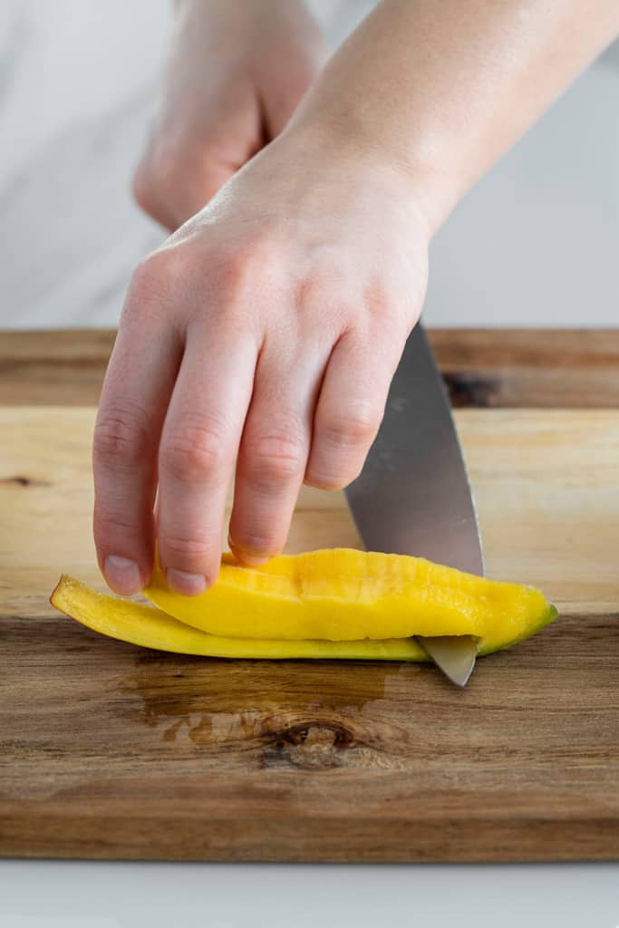 mango slice on cutting board with knife slicing through to remove flesh from skin