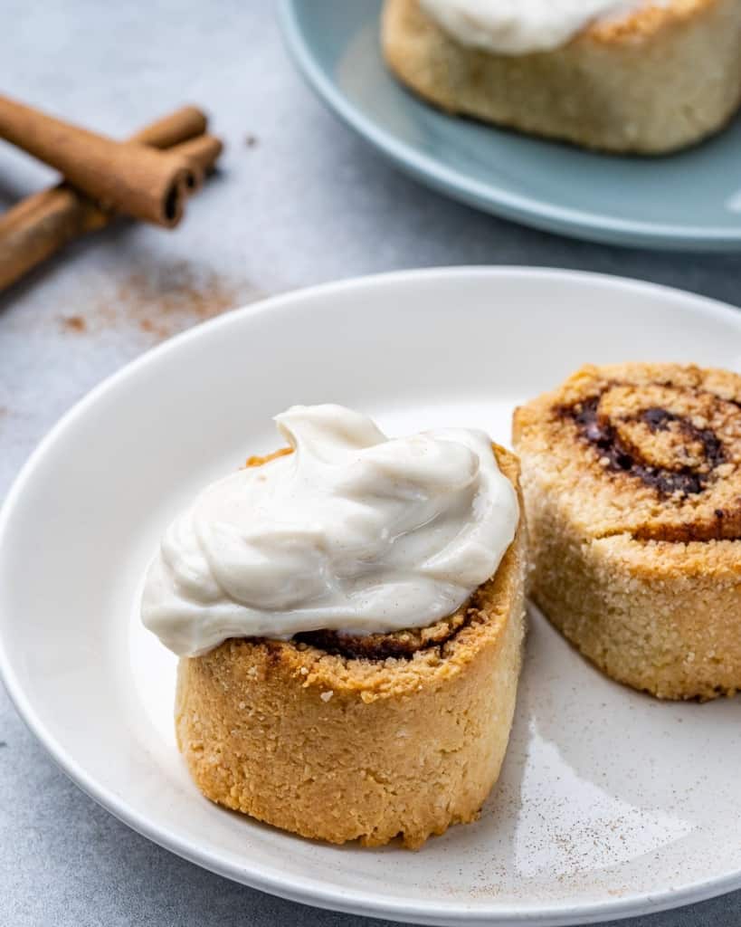 Two keto cinnamon rolls on white plate with cinnamon sticks in background