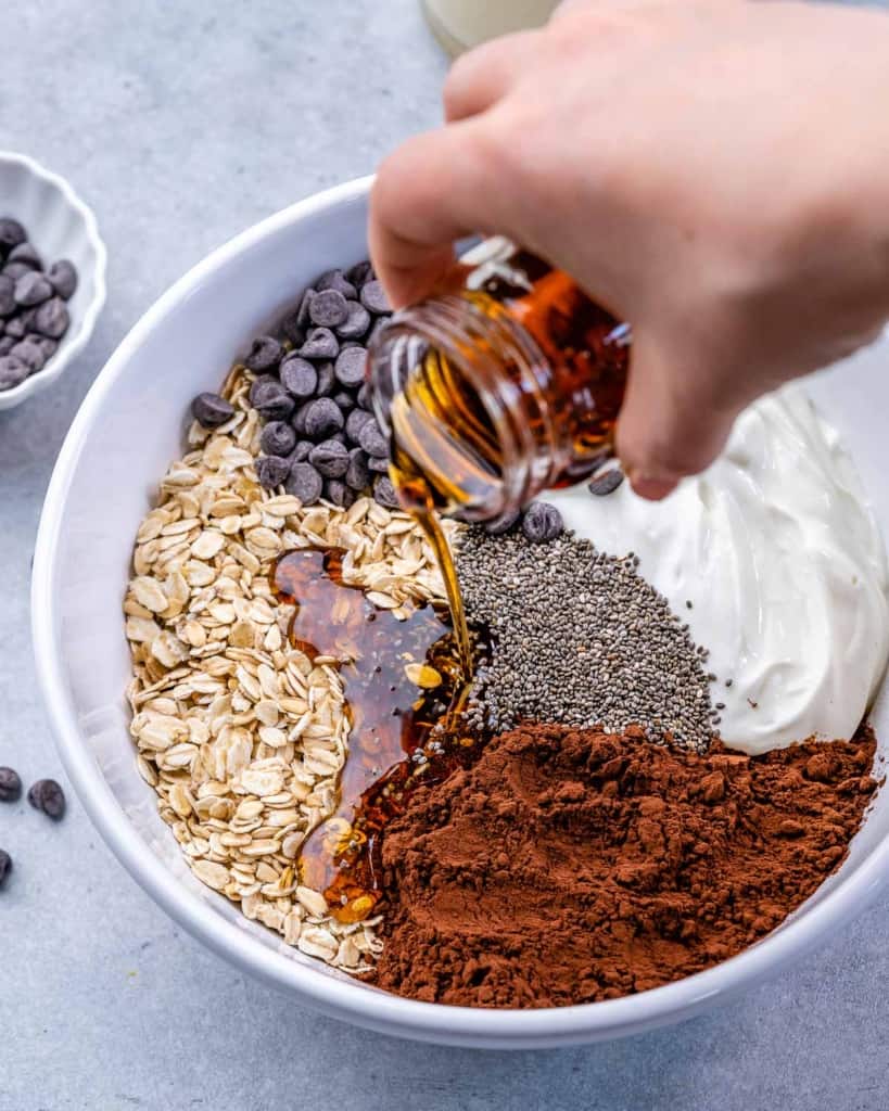 oats, chocolate chips, cacao powder, chia seeds, yogurt, and maple syrup being poured in white bowl 