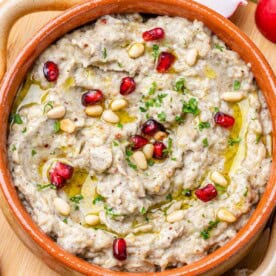 baba ganoush in a brown round bowl topped with pine nuts and pomegranate arils.