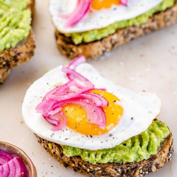 side shot of avocado toast topped with sunny side up eggs with purple pickled onions