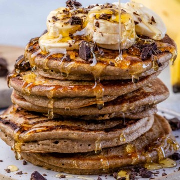 close up image of stacks of banana oatmeal pancakes topped with banana, shaved chocolate, and maple syrup drizzle