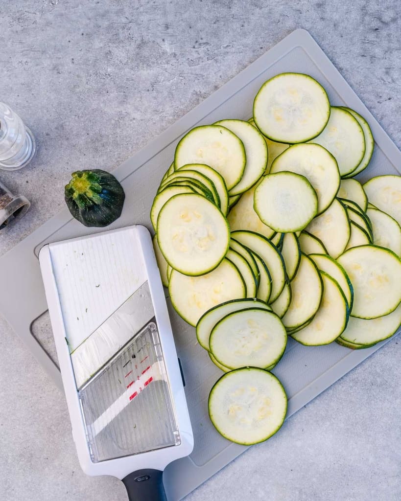 sliced zucchini next to mandolin on a cutting board